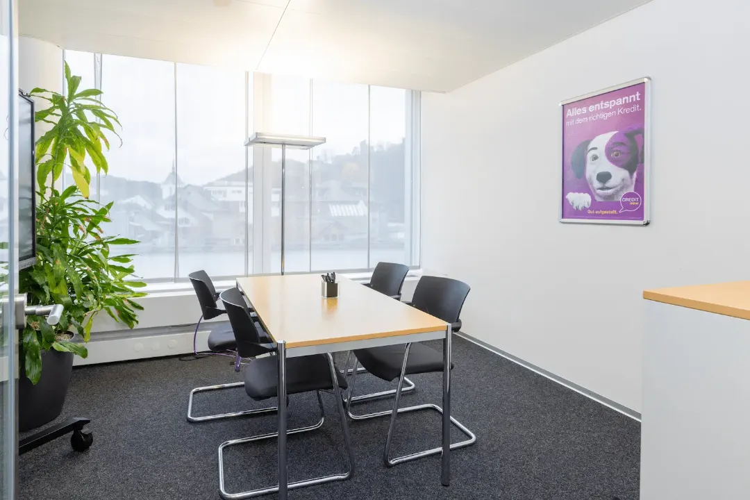 Besprechungsraum der BANK-now Filiale Baden mit Tisch, vier Stühlen, Pflanze und Poster.