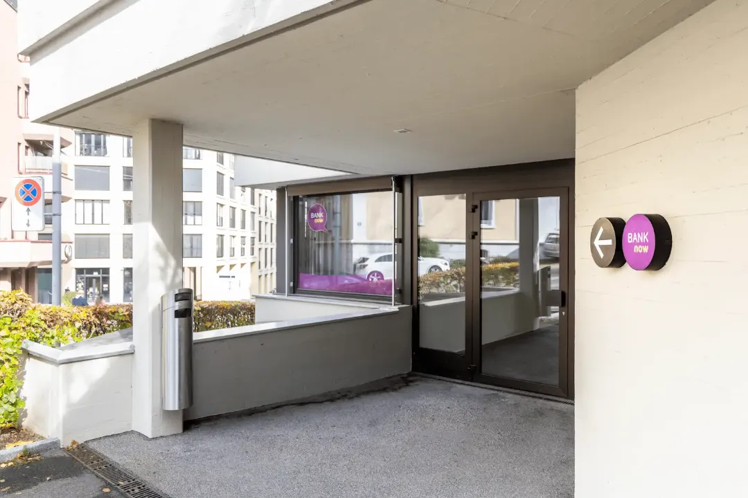 Entrance area of the BANK-now Zurich-Oerlikon branch under a canopy with glass doors and logo.
