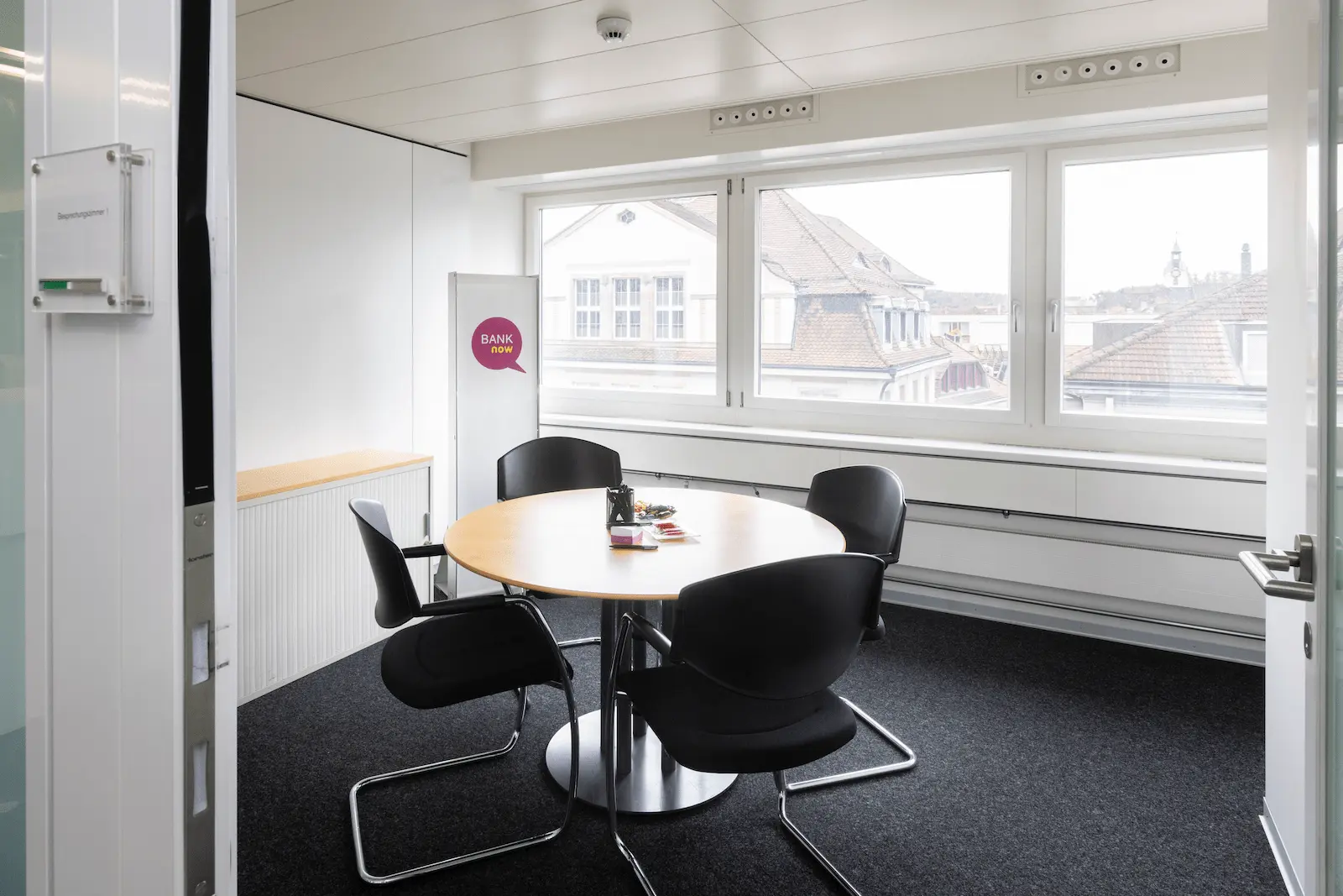 Besprechungsraum der BANK-now Filiale Olten mit rundem Tisch, Stühlen und grossem Fenster.