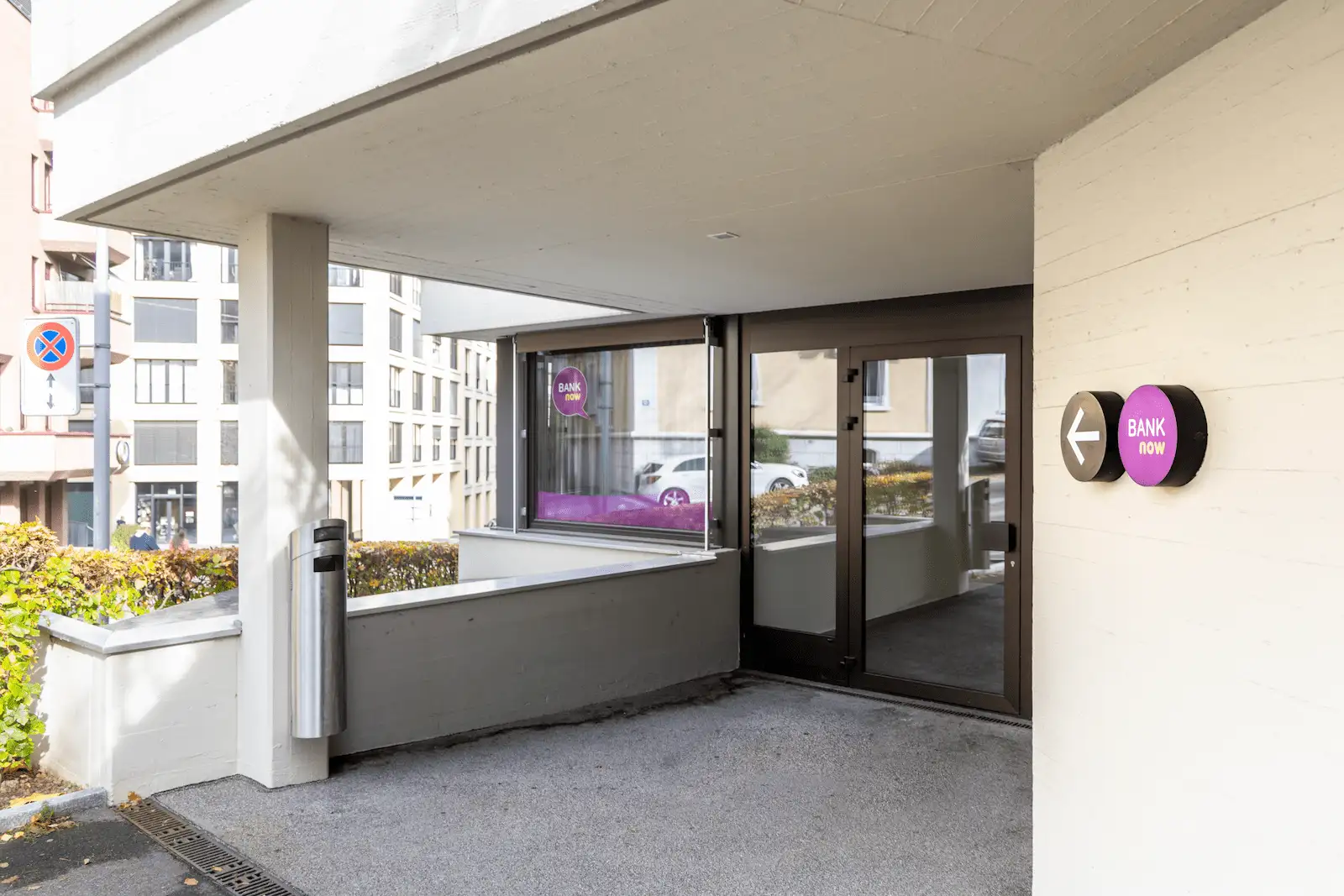 Entrance area of the BANK-now Zurich-Oerlikon branch under a canopy with glass doors and logo.