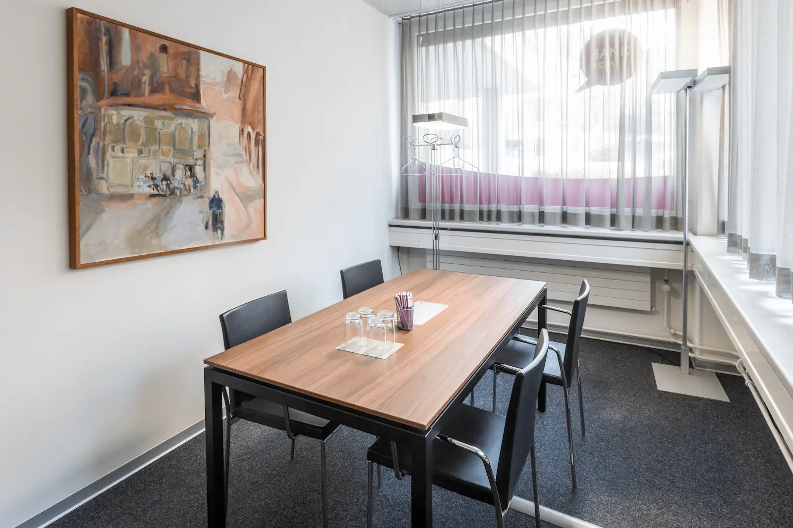 Meeting room of the BANK-now Zurich-Oerlikon branch with table, four chairs, wall painting and window front.